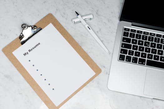 A clipboard with a resume beside a pen and laptop on a marble desk, ideal for business and job application themes.