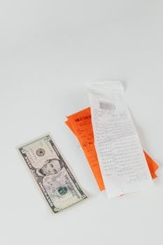 A crisp US dollar bill next to colorful shopping receipts on a clean white surface.